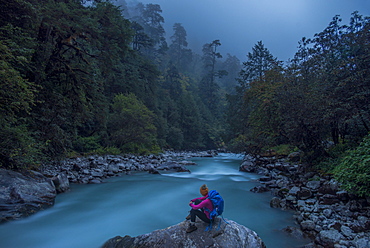 A woman takes a break from the trail and sits beside the Langtang Khola near the little village of Riverside on a misty evening, Nepal, Himalayas, Asia