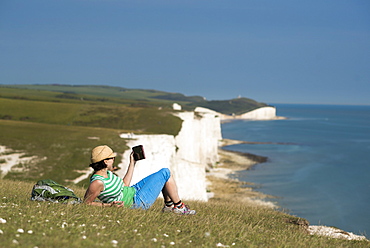 A woman reads her iPad on the cliffs with views of the Seven Sisters coastline in the distance, South Downs National Park, East Sussex, England, United Kingdom, Europe