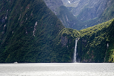 A sightseeing ship dwarfed by a tall waterfall in a fjord, South Island, New Zealand, Pacific