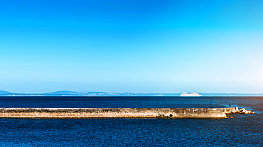 A single person stands on a long stone breakwater jutting out into the ocean with a large cruise ship in the distance, Estoril, Portugal, Europe