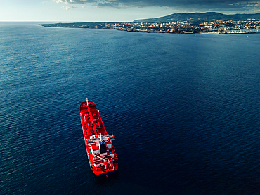 A large red cargo ship floats on the calm waters, close to the shoreline of a coastal city