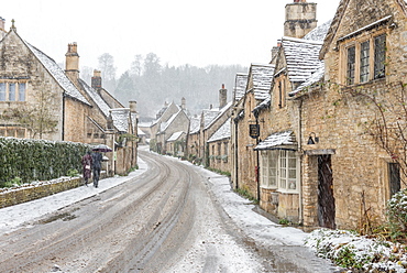 A couple walking up the hill in Castle Combe with an umbrella protecting them from the heavy snowfall, Wiltshire, England, United Kingdom, Europe