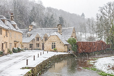 Snow covered houses by By Brook in Castle Combe with a dog enjoying a paddle, Wiltshire, England, United Kingdom, Europe