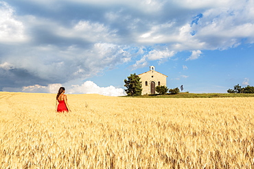 Woman in red dress admiring Notre-Dame-de-Sante chapel in a wheat field, Entrevennes, Alpes-de-Haute-Provence, Provence-Alpes-Cote d'Azur, France, Europe