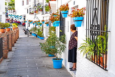 An elderly woman stands in front of a whitewashed house covered in blue plant pots in Mijas Pueblo, Andalusia, Spain, Europe