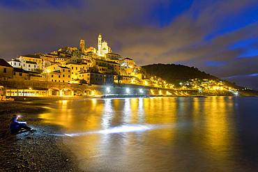 A tourist sitting on the beach observes the village at night, Cervo, Imperia province, Liguria, Italy, Europe