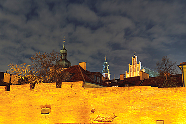 Night low angle view of illuminated defensive wall fortification in front of traditional low-rise roof-tiled houses and passing clouds above in the Old Town of Warsaw, Poland, Europe