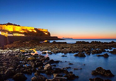 View from rocky shore to the illuminated Fortezza and distant Akrotiri Peninsula, sunset, Rethymno (Rethymnon), Crete, Greek Islands, Greece, Europe