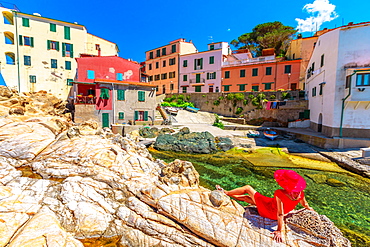 Woman in red dress sitting on cliffs at Marciana Marina in Borgo al Cotone with the famous houses of old village in bay of Marciana Marina promenade, Elba Island, Tuscany, Italy, Europe