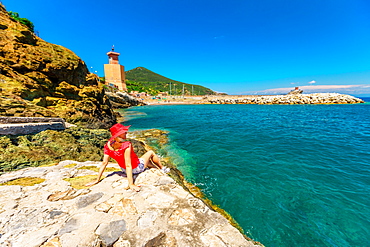 Happy woman in red hat resting on cliffs in Rio Marina beach, with clock tower in the background, Rio Marina, Elba island, Tuscany, Italy, Europe