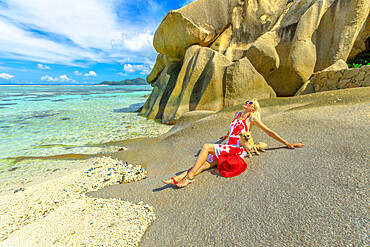 Tourist woman and cute dog sitting on beach at low tide, with granite rocks behind, at Anse Source d'Argent, La Digue, Seychelles, Indian Ocean, Africa