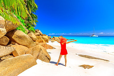 Tourist woman in red dress looking at turquoise sea and sailing boat and catamarans on horizon, Praslin Island, Seychelles, Indian Ocean, Africa