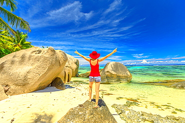 Tourist woman with raised arms in red hat standing on a boulder, Anse Source d'Argent, La Digue, Seychelles, Indian Ocean, Africa