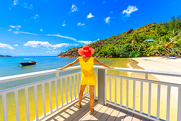 Woman in yellow dress on balcony of wooden jetty, looking at pristine white beach of Anse Gouvernement, near Cote d'Or Bay, in Praslin, Seychelles, Indian Ocean, Africa