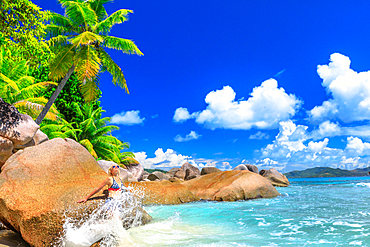 Tourist woman in splashing wave at Felicite Island, boulders and palm trees behind, La Digue, Seychelles, Indian Ocean, Africa