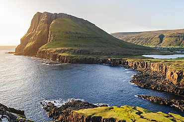 A panoramic view of a rugged coastal landscape of Suduroy in the Faroe Islands. The image captures a dramatic cliff face overlooking a narrow inlet, surrounded by the vast Atlantic Ocean.