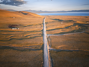 A long, dusty road stretches across the vast landscape of Song Kol Lake in Kyrgyzstan, leading towards the horizon and a distant body of water