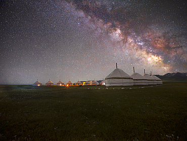 A row of white yurts is illuminated by the Milky Way, stretching across the night sky. The yurts are situated on a dark, grassy field in the SongKol Lake area of Kyrgyzstan