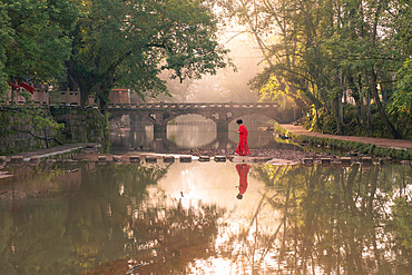 An ancient stone bridge in Shangli Town reflected in the river shrouded in morning mist and a women in red on a walkway on the river