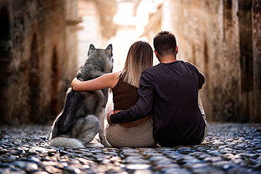 Back view of two young people and their beloved wolf dog sitting and hugging in an old town street staring at the sun, Piemonte, Italy, Europe