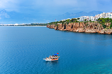 Antalya city view near the cliffs with a tourist boat on the sea with turquoise water and mountains in the background, in Turkey