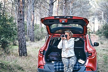 Young adult caucasian woman taking pictures close to a car parked in a forestry path in autumn. Travels concept. Ayegui, Navarre, Spain, Europe.