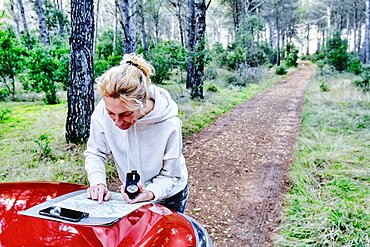 Young adult caucasian woman orienting herself with a map and a compass close to a car parked in a forestry path. Travels concept. Ayegui, Navarre, Spain, Europe.