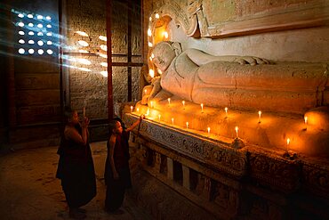 Two novice monks lighting candle by Buddhist statue inside temple, UNESCO, Bagan, Myanmar