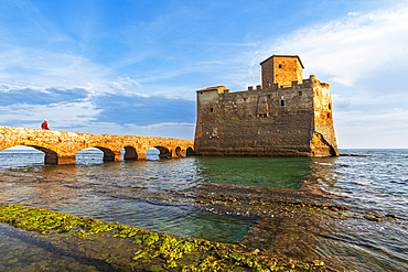 Man on the bridge walks to the fortified castle of Torre Astura in the water of Tyrrhenian Sea built on ruins of Roman villa, sunset time, Rome province, Latium (Lazio), Italy, Europe