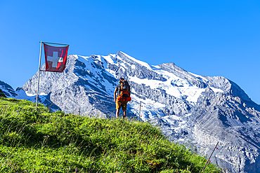 Rear view of a man with backpack hiking the Swiss Alps passing a Swiss flag flying, Oeschinensee, Kandersteg, Bern Canton, Switzerland, Europe