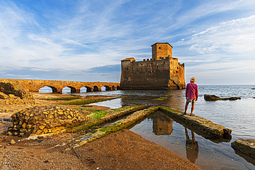 Man with hat stands on top of Roman ruin facing the medieval castle of Torre Astura rising from the sea, Nettuno municipality, province of Rome, Tyrrhenian sea, Latium (Lazio), Italy, Europe