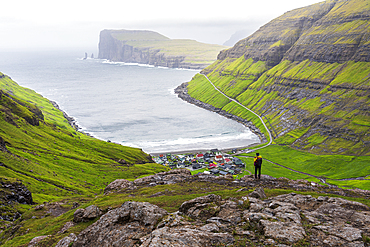 One man on top of a rock admiring the village of Tjornuvik in the misty weather, Sunda municipality, Streymoy island, Faroe islands, Denmark, Europe