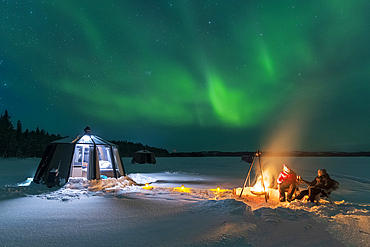 Night view of two people enjoyng dinner outside around a campfire close to the illuminated glass igloo with Northern Lights (Aurora Borealis) dancing in the sky, Jokkmokk, Norrbotten, Swedish Lapland, Sweden, Scandinavia, Europe