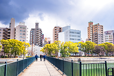 Pedestrian walkway over the river in Hiroshima leading to a cityscape of mid-rise buildings and trees under dramatic clouds