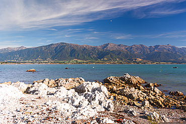Rocky shoreline with dramatic mountain backdrop under blue sky at Kaikōura Peninsula, South Island, New Zealand
