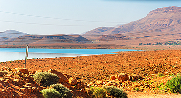 Arid vegetation and sunlit hills meet the blue expanse of Barrage Al-Hassan Addakhil in Parc National Al Hassan Addakhil