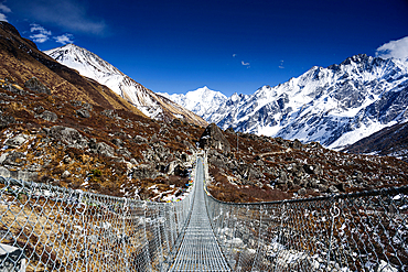 Suspension bridge leading into the snowy expanse of Langtang Valley with clear views of Gangchempo’s striking summit under crisp sunlight