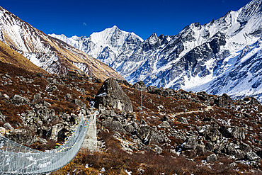 Suspension bridge crossing rocky terrain in Langtang Valley with panoramic views of Gangchempo and surrounding Himalayan peaks under bright blue skies
