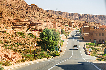 Paved desert road winding through the sandstone cliffs of Ziz Valley, Morocco, with small buildings scattered along the slope