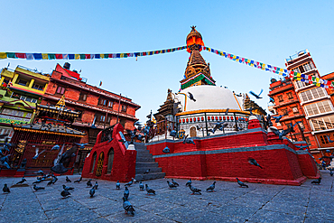 Kathi Swoyambhu Shree Gha Stupa in Kathmandu with pigeons, prayer flags, and vibrant surrounding buildings at sunset