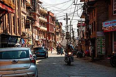 Motorcyclists and pedestrians navigating a sunlit narrow street lined with traditional and modern buildings in Bhaktapur