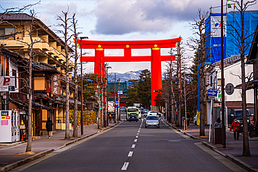 Urban street scene in Kyoto with a bright red torii gate spanning the road, surrounded by buildings and leafless winter trees