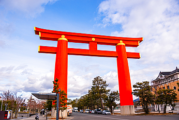 Urban street scene in Kyoto with a bright red torii gate spanning the road, surrounded by buildings and leafless winter trees
