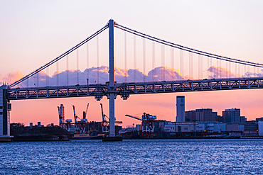 Industrial skyline and cargo cranes beneath the Rainbow Bridge at sunset, viewed from the waterfront in Tokyo Bay