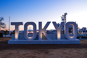 Large illuminated TOKYO sign at sunset with Rainbow Bridge and city skyline in the background at Odaiba Seaside Park, Tokyo, Japan