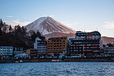 Snow-capped Mount Fuji rising above lakeside buildings at dusk in Kawaguchiko, Japan, viewed across the tranquil waters of Lake Kawaguchi