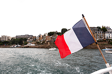 French flag flutters on the stern of a boat departing Dinard with the breton architecture visible across the water in Brittany France