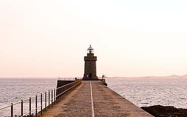 A stone pier leads to a lighthouse at the entrance of Saint Peter Port Harbour in Guernsey Channel Islands during a soft pastel sunrise