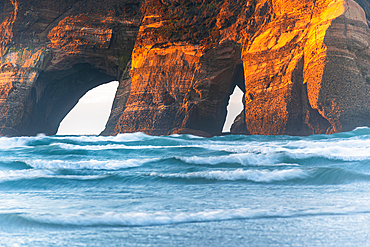 Sunlit cliffs of the Archway Islands glow above waves at Wharariki Beach, Golden Bay, New Zealand.