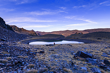 A hiker walking beside a reflective alpine lake in the high mountains of Cerro del Caballo, Sierra Nevada National Park, Andalusia, Spain.